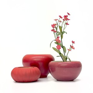 May include: Three red ceramic pots of varying sizes, with a sprig of pink flowers in the largest pot.