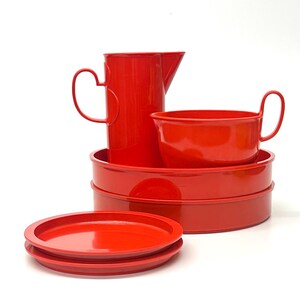 May include: A set of bright red kitchenware, including a pitcher, a bowl with a handle, two plates, and two trays. The pitcher has a curved handle. The items are arranged on a white background.