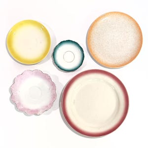 May include: A collection of five ceramic plates and saucers arranged on a white surface. The set includes a yellow plate, a small teal scalloped saucer, a speckled orange plate, a pink scalloped saucer, and a large plate with a deep red rim.