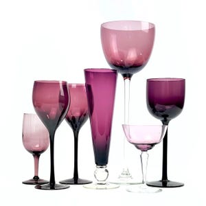 May include: A collection of six assorted drinking glasses in varying shades of purple. The set includes several stemmed wine glasses, a tall slender flute, and a wide shallow coupe glass, all featuring clear or dark purple stems and bases.