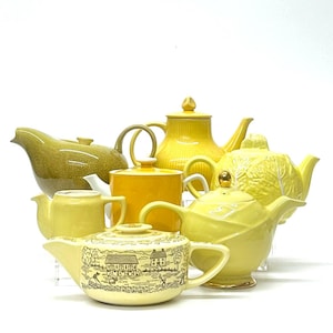 May include: A collection of yellow teapots in various shapes and designs. One teapot has a landscape scene, another resembles a cabbage, and others have ribbed or textured surfaces. The teapots are arranged on a white surface.