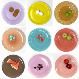 May include: A collection of nine round ceramic plates in various colors including lavender, yellow, tan, peach, turquoise, coral, brown, light pink, and rose pink, each displaying different food items like cookies, eggs, fruit, and berries.