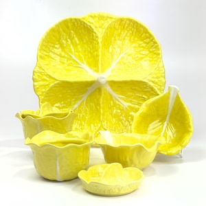 May include: A set of bright yellow ceramic tableware featuring a large leaf-shaped platter, three deep bowls with ruffled edges, and a small citrus juicer. Each piece displays a textured surface mimicking the veins and structure of a leaf.