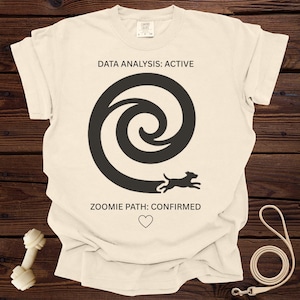 May include: Cream-colored t-shirt with a black spiral design and a running dog silhouette. Text above reads "DATA ANALYSIS: ACTIVE," and below, "ZOOMIE PATH: CONFIRMED" with a heart symbol. A dog toy and leash are visible.
