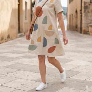 May include: A beige linen dress with short sleeves and a knee-length hem. The dress features colorful geometric shapes in orange, blue, yellow, and green. The person is wearing white sneakers and a brown crossbody bag. The dress has a relaxed fit.