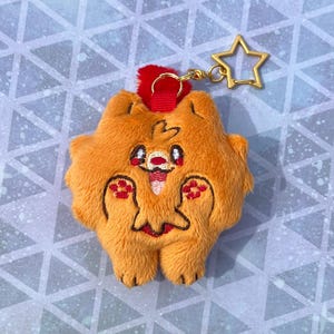 May include: Orange plush keychain in the shape of a cartoon animal, featuring embroidered details in red, black, and white. The keychain has a red loop and a gold star-shaped clasp.