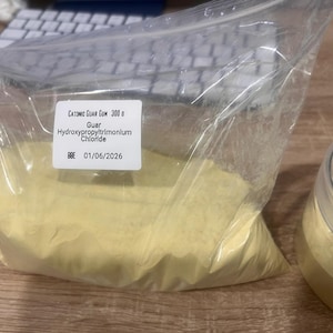 Puede incluir: Una bolsa de plástico transparente llena de polvo amarillo, etiquetada como "Cationic Guar Gum 300 g". Un tarro transparente con tapa contiene un polvo amarillo similar. La etiqueta incluye la fecha de caducidad 01/06/2026.