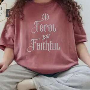 May include: Crimson-colored t-shirt with the text "Feral but Faithful" in a decorative font. The shirt has short sleeves and a relaxed fit. A logo with a butterfly and the words "Christ Wheel" is visible in the upper left corner.