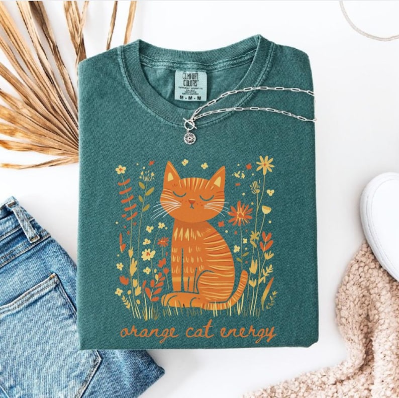 Pu&ograve; includere: Felpa color verde acqua con una grafica di un gatto arancione circondato da fiori. Il gatto ha gli occhi chiusi ed &egrave; seduto. Il testo "orange cat energy" &egrave; sotto il gatto. Una collana d'argento con un ciondolo &egrave; sulla maglietta.