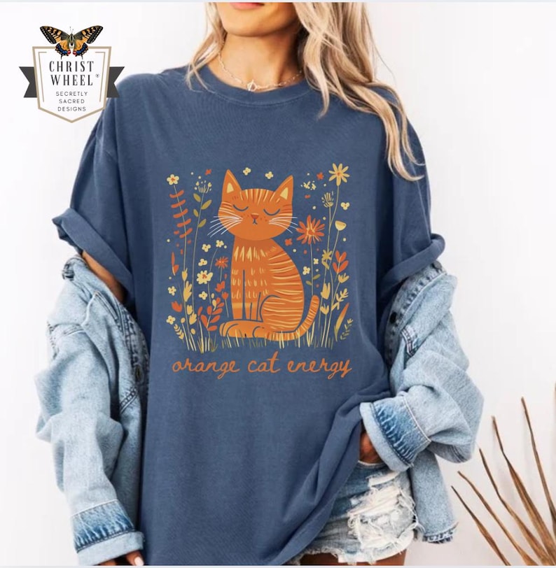 Pu&ograve; includere: T-shirt blu navy con un gatto arancione e fiori. Il testo "orange cat energy" &egrave; stampato sotto. La maglietta ha maniche arrotolate e una vestibilit&agrave; ampia.