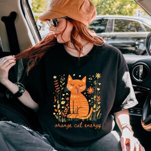 Pu&ograve; includere: T-shirt nero con un'illustrazione di un gatto arancione e la scritta "orange cat energy". Il gatto &egrave; circondato da elementi floreali gialli e arancioni. La persona che indossa la maglietta indossa un cappello di velluto a coste e occhiali da sole.