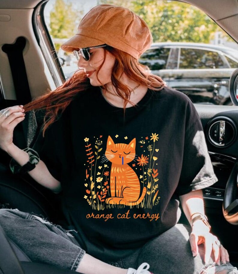 Pu&ograve; includere: T-shirt nero con un'illustrazione di un gatto arancione e la scritta "orange cat energy". Il gatto &egrave; circondato da elementi floreali gialli e arancioni. La persona che indossa la maglietta indossa un cappello di velluto a coste e occhiali da sole.