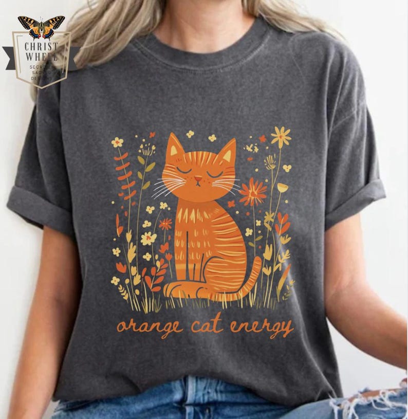 May include: A gray t-shirt with an orange cat sitting in a field of flowers. The cat has closed eyes and is surrounded by yellow, orange, and green flowers. The text 'orange cat energy' is printed below the cat.