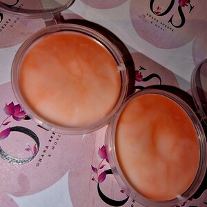 Könnte beinhalten: Zwei transparente Plastikbehälter mit Deckeln, jeder mit einem pfirsichfarbenen Lippenbalsam gefüllt. Die Behälter befinden sich auf einer weißen Oberfläche mit pinkfarbenen und weißen, floralen Aufklebern, auf denen "Ohana Scents & More" steht.