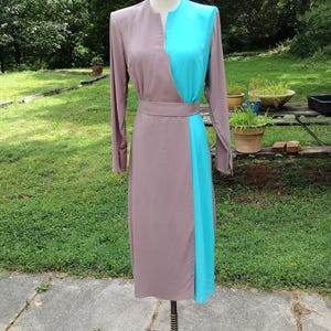 Op de afbeelding: Een lange mouwen jurk met een tweekleurig ontwerp. De bovenste helft van de jurk is turquoise blauw, en de onderste helft is lichtbruin. De jurk is op een mannequin en wordt getoond tegen een groene gras achtergrond.