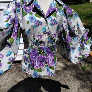 May include: A long-sleeved button-down shirt featuring a white base with a vibrant floral pattern in shades of purple, blue, and green. The garment has a pointed collar, a front button closure, and voluminous, gathered sleeves at the shoulders.