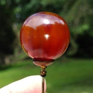 May include: A stick pin featuring a smooth, spherical, translucent deep amber-colored bead. The bead is mounted on a small, metallic, gold-toned cup-shaped setting that connects to a thin, straight, metallic pin shaft extending downward.