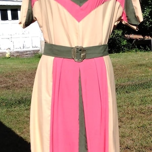 May include: A beige dress featuring a V-neckline, short sleeves, and a color-blocked chevron pattern in olive green and bright pink across the chest. The dress includes an olive green belt with a rectangular buckle and a pink pleated skirt panel.