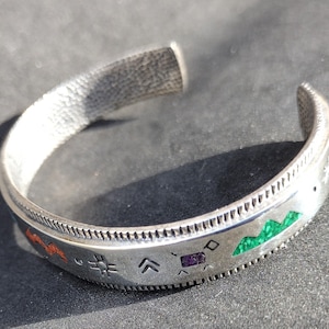 May include: Silver cuff bracelet with etched geometric designs and colourful inlaid accents. The bracelet features red, green, and purple details, with a textured finish. A stylish accessory, suitable for any occasion.