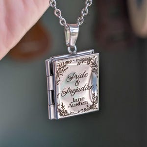 Peut inclure: Un médaillon en métal argenté en forme de livre miniature, suspendu à une chaîne à maillons assortie. La couverture avant présente une bordure florale gravée et le texte Pride & Prejudice de Jane Austen dans une police script et serif.