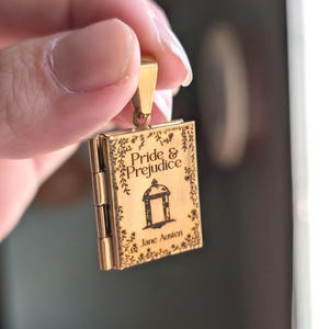 Könnte beinhalten: Ein goldfarbenes Metallmedaillon in Form eines Miniaturbuchs. Das Cover zeigt den eingravierten Text "Pride & Prejudice" und "Jane Austen" sowie eine zentrale Abbildung eines Pavillons, umrahmt von einem zarten floralen Rankenmuster.