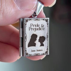 May include: A silver-toned metal book-shaped locket pendant featuring an engraved cover with the text Pride & Prejudice and Jane Austen, accompanied by two dark silhouettes of figures facing each other, framed by decorative scrollwork borders.