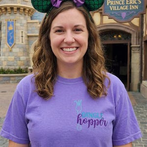 May include: A woman wearing a purple t-shirt with the text "Dinglehopper" and a pair of green and purple mouse ears with a bow. The background features a castle and a restaurant sign that reads "Prince Eric's Village Inn."