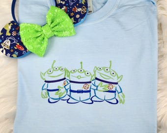 Puede incluir: Camiseta azul claro con una imagen bordada de tres extraterrestres verdes de la película Toy Story. Los extraterrestres llevan trajes espaciales azules y tienen tres ojos cada uno.