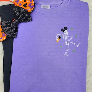 May include: A purple Comfort Colors sweatshirt with a skeleton wearing Mickey Mouse ears and holding an ice cream cone. The design is embroidered in white and green. A black shirt and Halloween-themed mouse ears are also in the image.