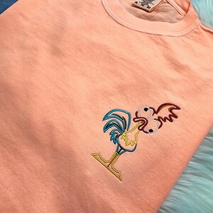 May include: Peach coloured t-shirt with an embroidered cartoon rooster with large eyes. The rooster is outlined in blue, yellow, and red.