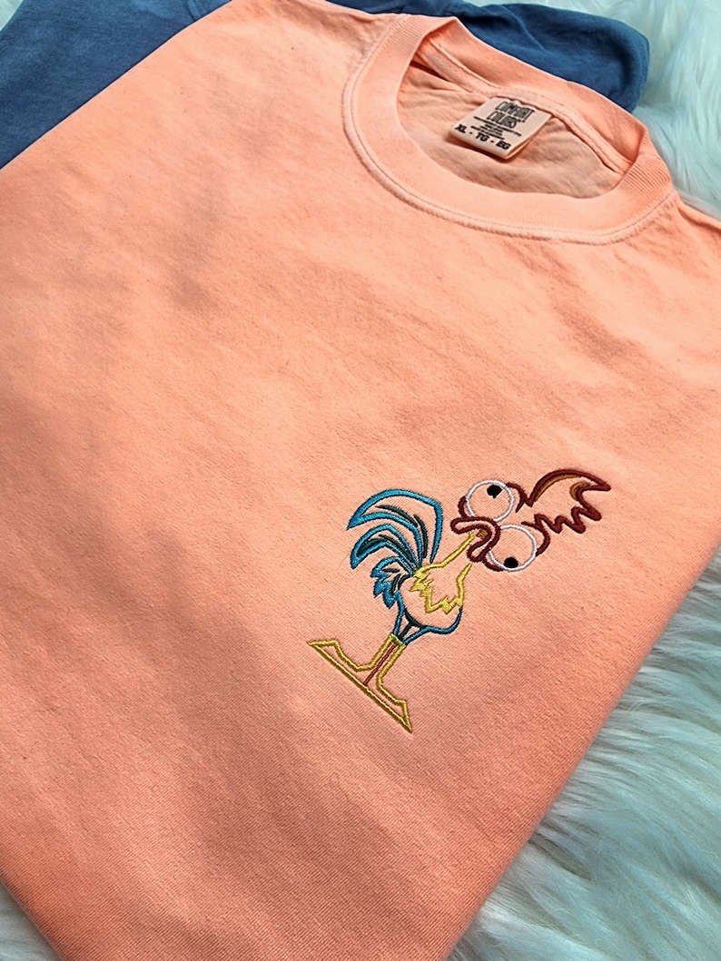 May include: Peach coloured t-shirt with an embroidered cartoon rooster with large eyes. The rooster is outlined in blue, yellow, and red.