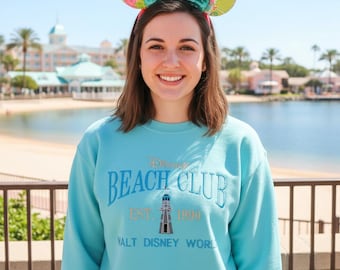 May include: A light blue sweatshirt with the text "Beach Club" and a lighthouse graphic. The sweatshirt is from Walt Disney World, established in 1990. The person is wearing mouse ears with a floral pattern and a teal bow.