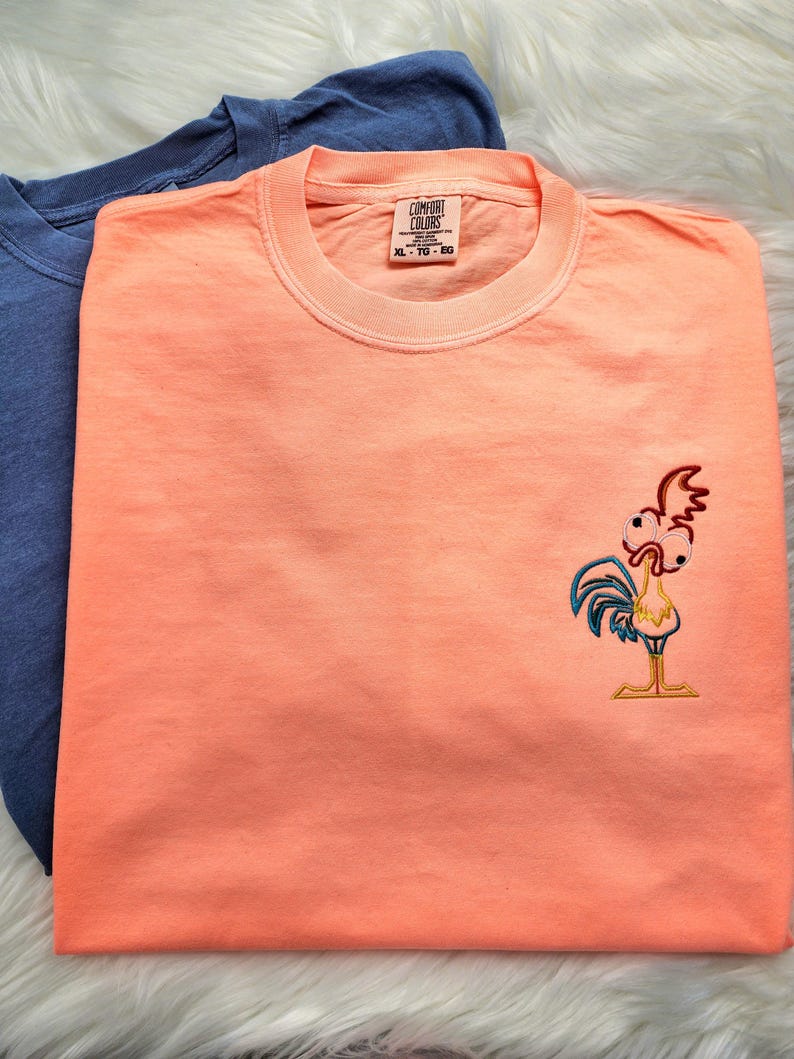 May include: A peach coloured t-shirt with a cartoon rooster embroidered on the front. The shirt has a tag that says "Comfort Colours" and "XL - 1G - 5G".