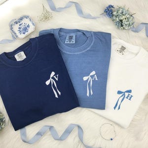 May include: Three crewneck t-shirts in navy blue, light blue, and white. Each shirt features a small embroidered bow graphic on the left chest. The navy and light blue shirts have white bows with letters V and A, while the white shirt has a blue bow with letter H.