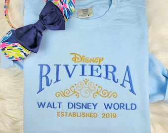 May include: Light blue sweatshirt with gold and blue embroidered text that reads "Disney Riviera Walt Disney World Established 2019".