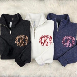 May include: Three quarter-zip pullovers in charcoal gray, white, and navy blue. Each garment features a silver metal zipper at the collar and a large, decorative embroidered monogram on the chest in peach, red, and light pink thread respectively.