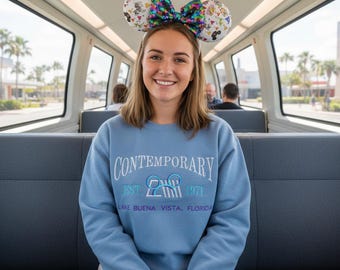 May include: A woman wearing a light blue sweatshirt with the word "Contemporary" and the text "EST. 1971 Lake Buena Vista, Florida". She is also wearing a headband with Mickey Mouse ears and a sequined bow. The background shows a train interior.
