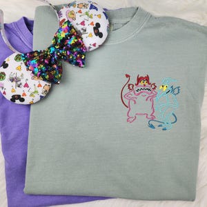 May include: A light green t-shirt with an embroidered image of two cartoon characters. The characters are a pink devil with red horns and a blue devil with wings. The t-shirt is paired with a purple t-shirt and a sparkly headband with a Disney theme.