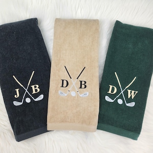 May include: Three golf towels in black, tan, and green. Each towel features embroidered golf clubs, a golf ball, and initials. The black towel has "JXB", the tan towel has "DB", and the green towel has "DXW".