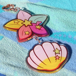 May include: A colorful flower charm with a pink, yellow, and green design and a seashell charm with a pink and yellow design. Both charms have gold stars and are attached to a silver key ring.