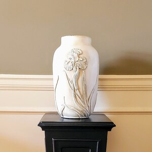 May include: A white ceramic vase with an embossed iris design sits on a black pedestal stand.