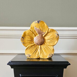 May include: A yellow ceramic flower with brown accents sits on a black pedestal. The flower has a textured surface and a raised center.