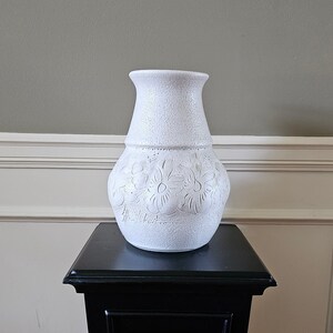 May include: White ceramic vase with a textured surface and floral design. The vase is sitting on a black pedestal.