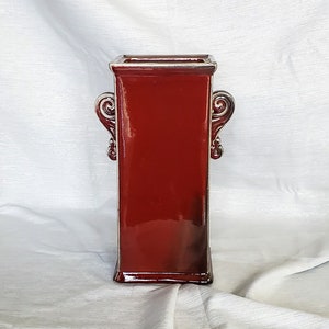 May include: A red ceramic vase with ornate silver handles. The vase has a square shape and a glossy finish.