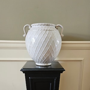 May include: A white ceramic vase with a diamond pattern and two handles sits on a black pedestal.