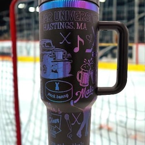 May include: A matte black insulated tumbler with a handle and a black lid, featuring iridescent purple and blue graphics including a jeep, hockey sticks, beer mugs, a microphone, and text reading "Hastings, MA", "puck bunny", and "you and suddenly know you needed".