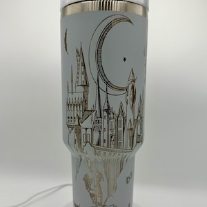 May include: A light blue insulated tumbler with a white lid and a gold-colored design. The design features a castle, a crescent moon, stars, and bats. The tumbler is approximately 7 inches tall.