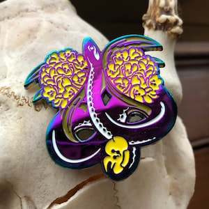 Puede incluir: Un colorido pin de esmalte con un diseño de pulpo con estructuras amarillas similares a cerebros y una calavera. El pin tiene un acabado púrpura e iridiscente con detalles en blanco y amarillo. El pulpo tiene múltiples tentáculos.