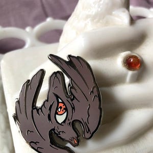 Puede incluir: Un pin de esmalte plateado con una ilustración detallada de un pájaro con un gran ojo naranja. El pájaro está representado con las alas extendidas, y el pin tiene un esquema de color gris oscuro.