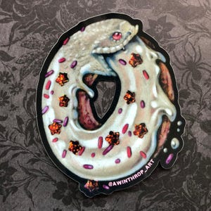 May include: A white donut with pink and orange sprinkles and a white gecko wrapped around it. The donut has a brown center and the gecko has a red eye. The sticker has the text "@AWINTHROP_ART" on the bottom.
